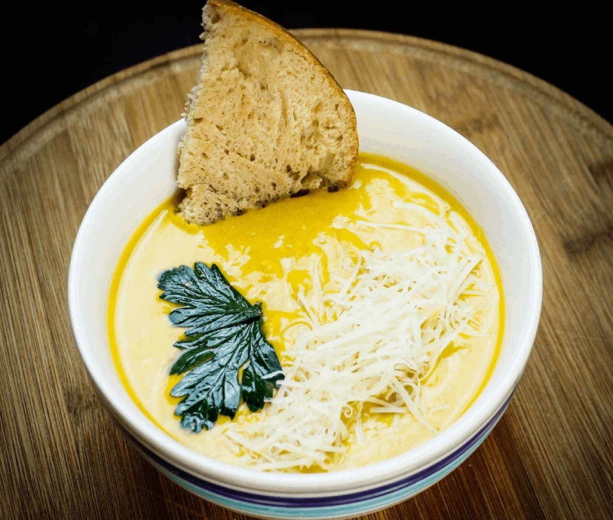 Biała misa z kremową żółtą zupą serową posypaną startym serem i ozdobiona natką pietruszki, obok kawałek świeżego chleba na drewnianej desce